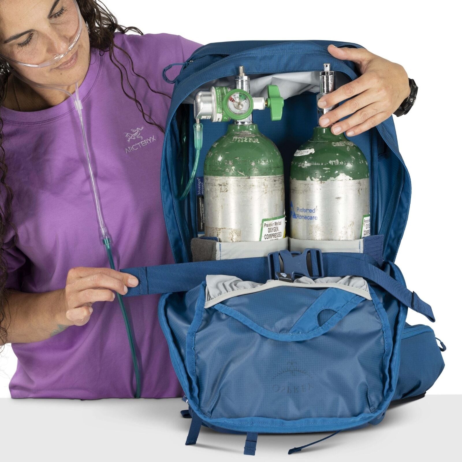A person in a purple shirt and oxygen tube inspects a blue backpack, revealing two green oxygen tanks inside. The setting is clinical and practical.