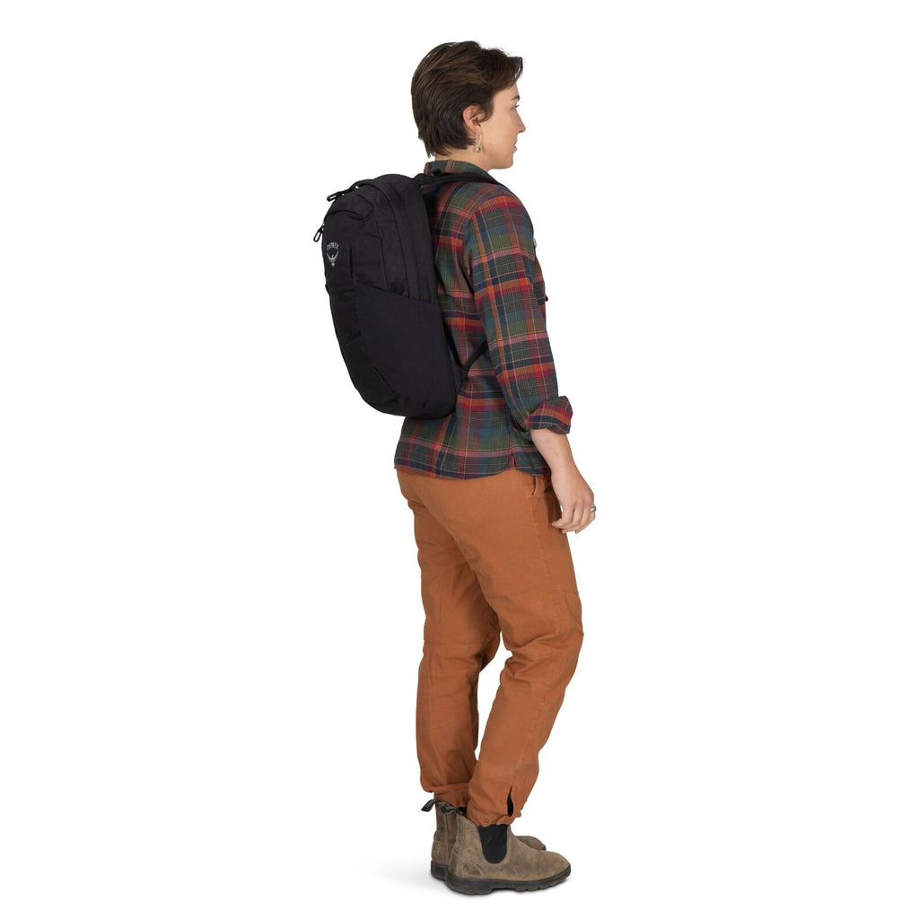Person dressed casually in a plaid shirt and brown pants, standing sideways with a black backpack. The background is plain white, conveying a neutral tone.