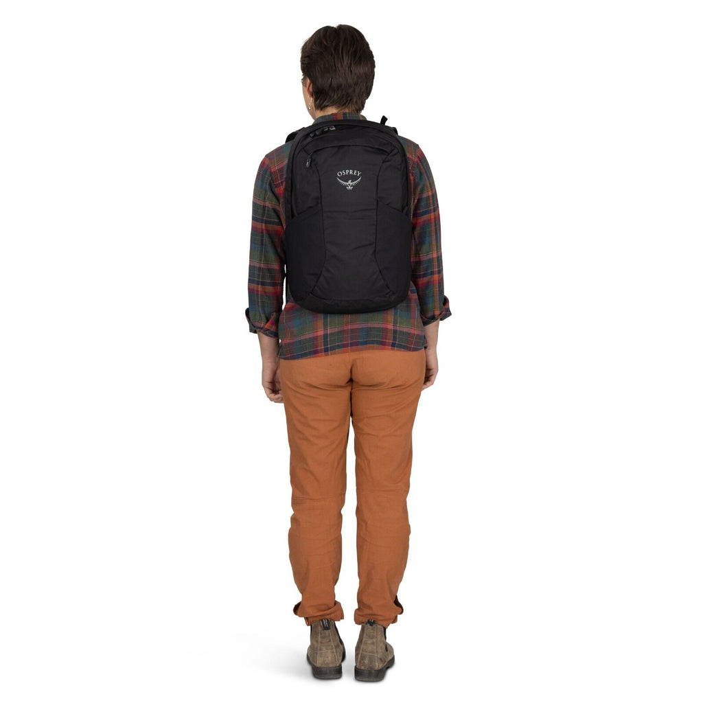 Person in plaid shirt and orange pants stands facing away, wearing a black Osprey backpack. The setting is neutral, conveying a casual tone.