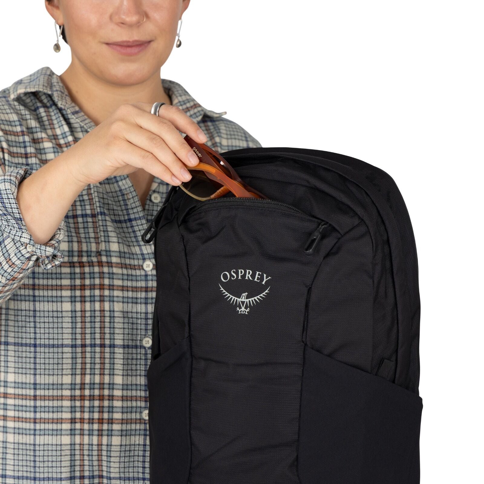 A person in a plaid shirt inserts sunglasses into the top pocket of a black Osprey backpack. The scene conveys organization and practicality.