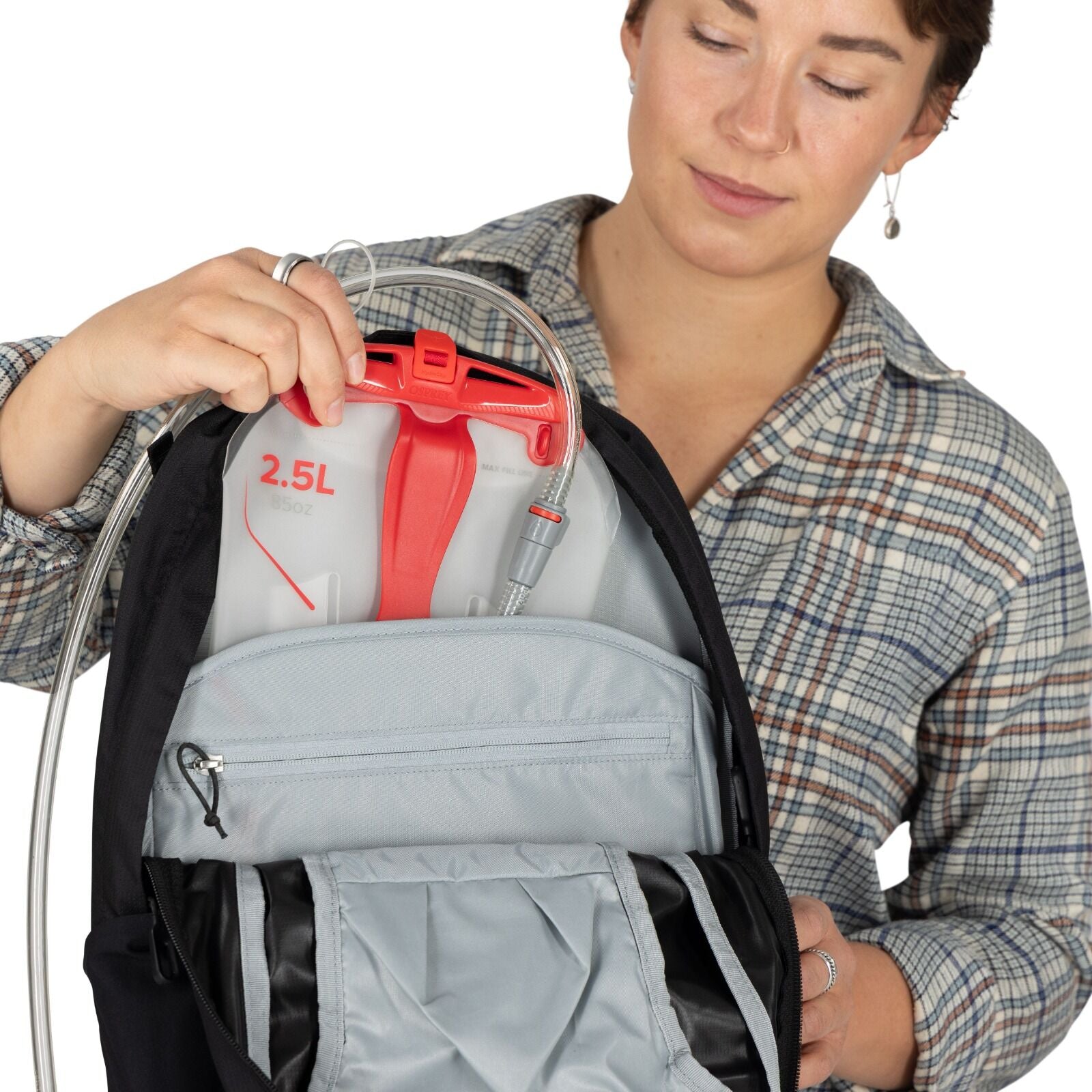 A person holds a backpack with a 2.5L hydration bladder inside. The bladder has a red handle and clear tube. The person wears a plaid shirt.