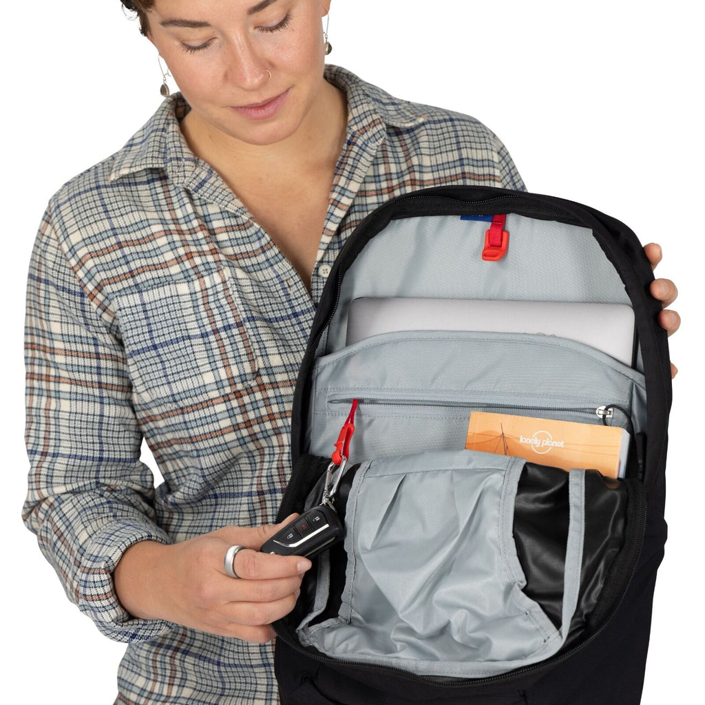 A person in a plaid shirt opens a backpack, revealing organized compartments and a travel guide. They hold a car key, suggesting travel or preparation.