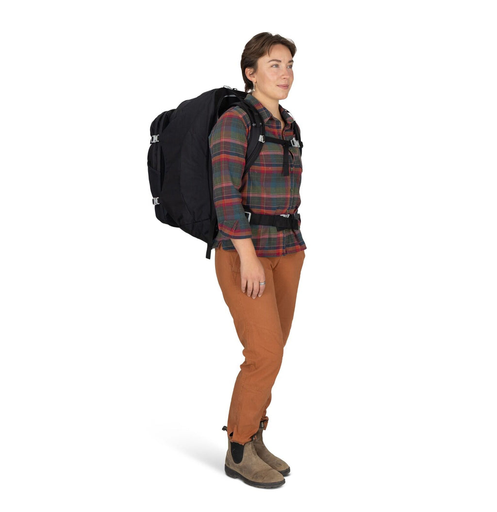 A person stands side view, wearing a large black backpack, plaid shirt, brown pants, and boots. The expression is calm and prepared, suggesting readiness for travel.