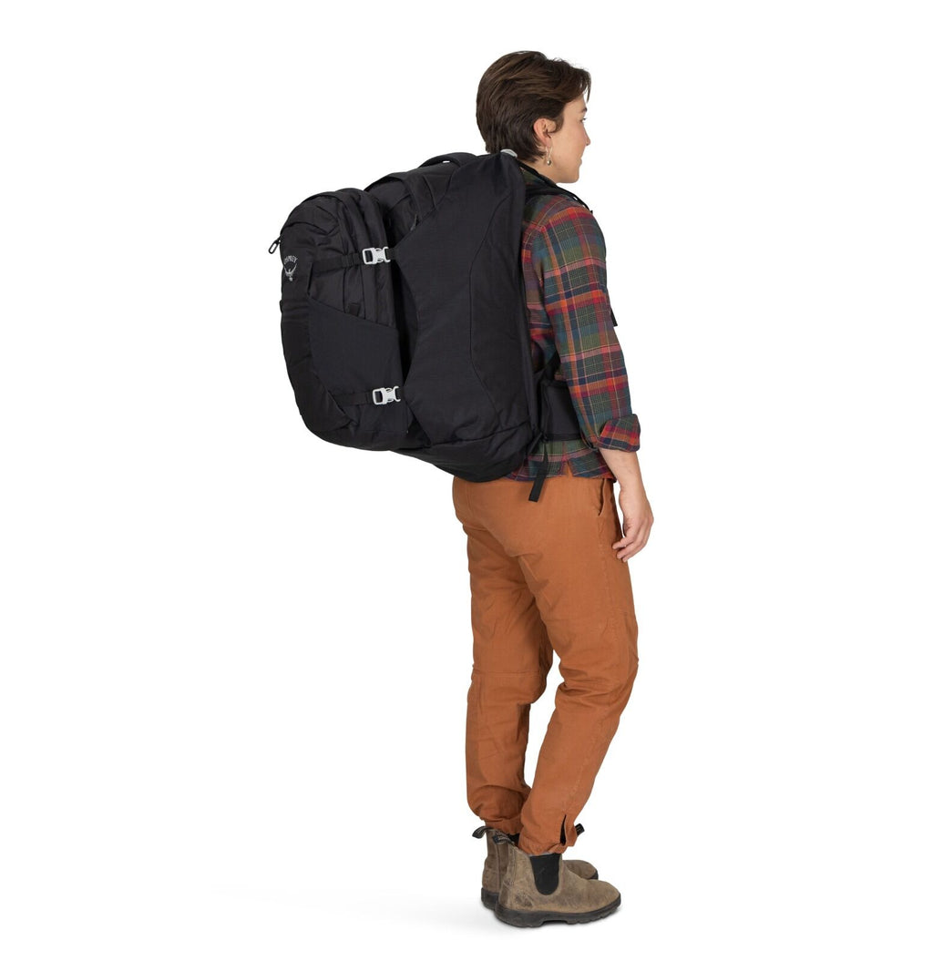 A person wearing a large black backpack, dressed in a plaid shirt, brown pants, and ankle boots, stands sideways against a white background.