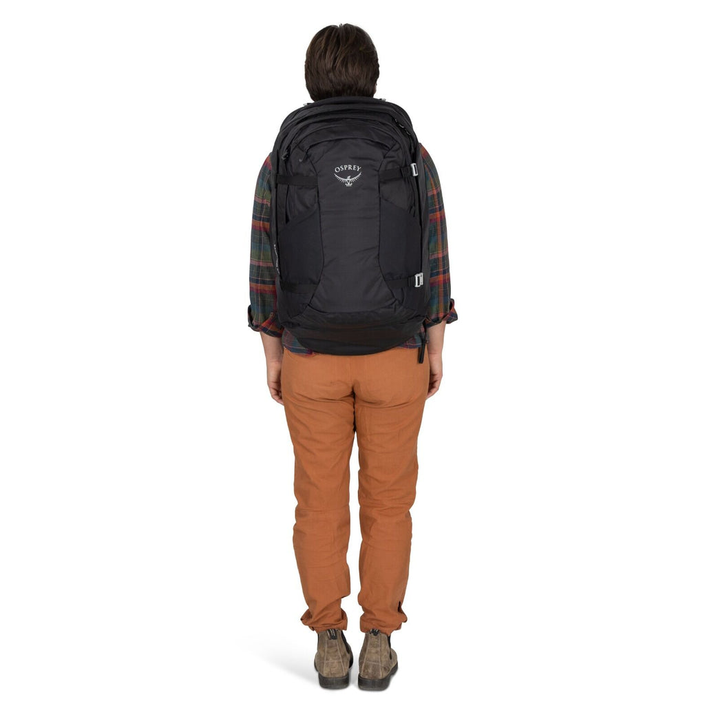 A person wearing a large black backpack, dressed in a plaid shirt, brown pants, and ankle boots, stands sideways against a white background.