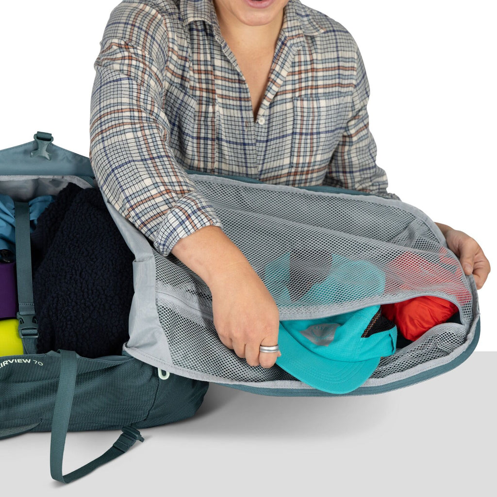 Person in a plaid shirt packs a teal and red clothing into a mesh compartment of a teal duffel bag. The scene conveys organization and travel readiness.