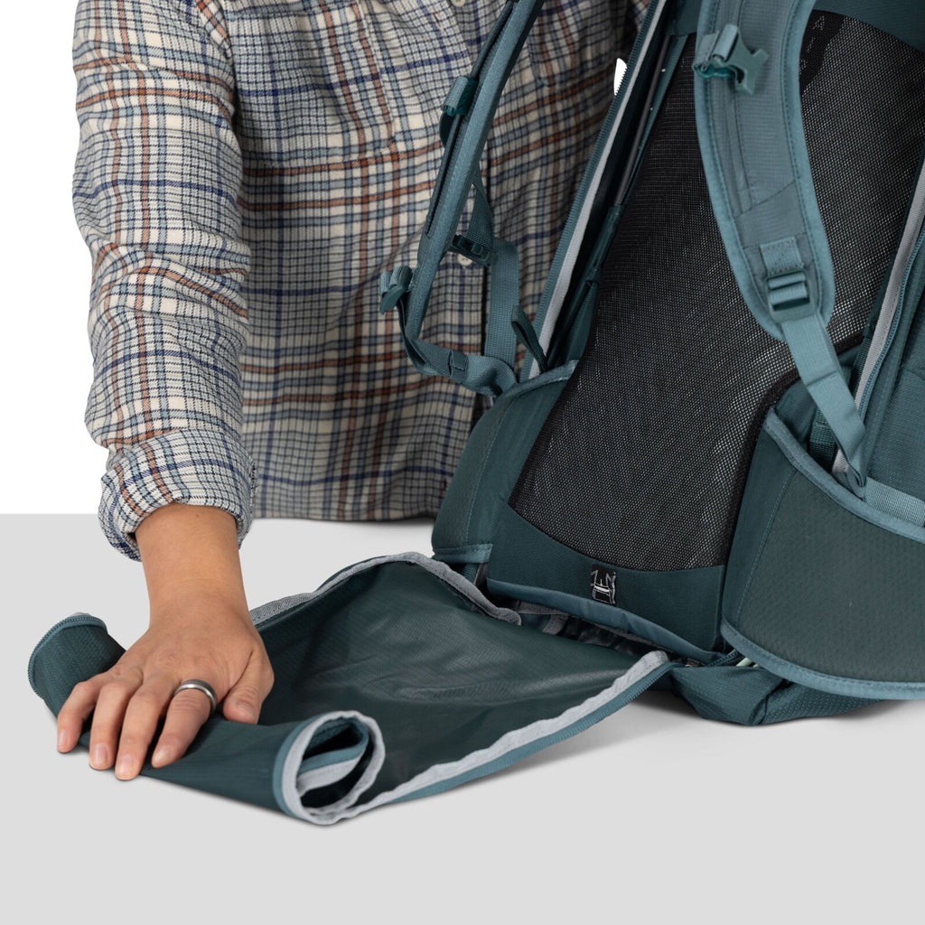 A person in a plaid shirt adjusts a teal backpack, focusing on rolling out a detachable section. The scene conveys preparation and organization.