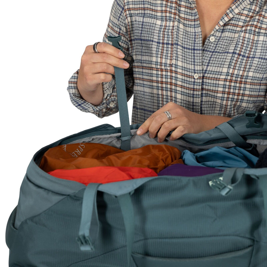A person in a plaid shirt opens a teal duffel bag filled with colorful clothes, conveying a sense of travel or preparation. Hands grasp a strap.