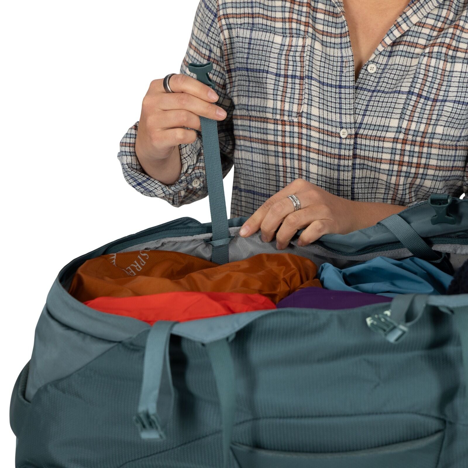 A person in a plaid shirt opens a teal duffel bag filled with colorful clothes, conveying a sense of travel or preparation. Hands grasp a strap.