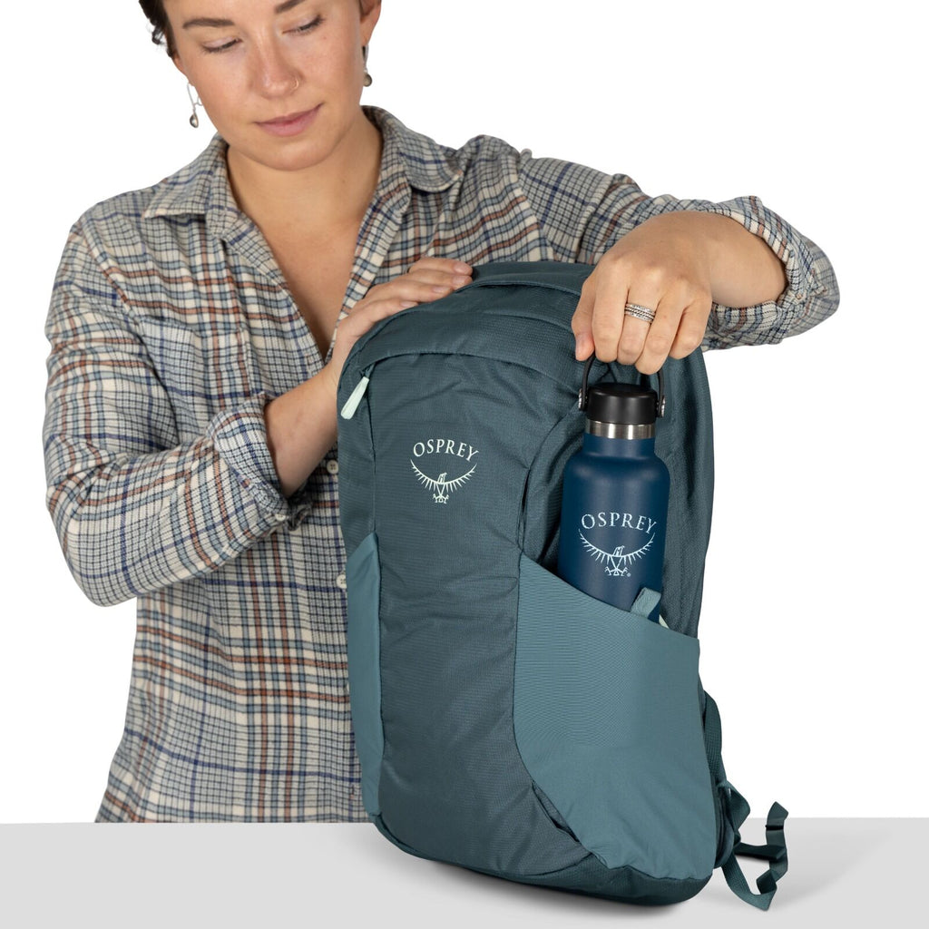 A person in a plaid shirt places a blue water bottle into a side pocket of a teal Osprey backpack. The setting is neutral and casual.