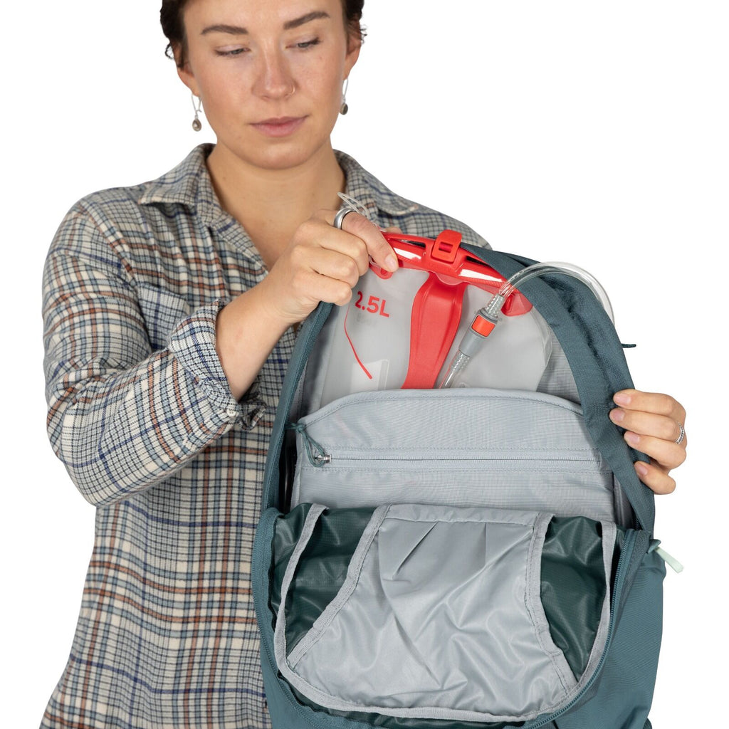 A person in a plaid shirt is holding open a blue backpack, showing a gray 2.5L hydration reservoir with a red cap and attached tube inside.