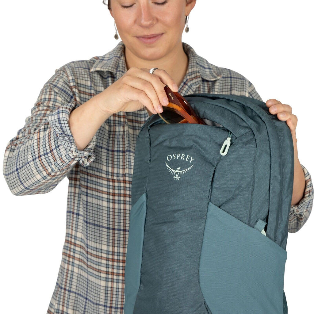A person in a plaid shirt places sunglasses into a blue Osprey backpack with a subtle smile, suggesting readiness for a journey.