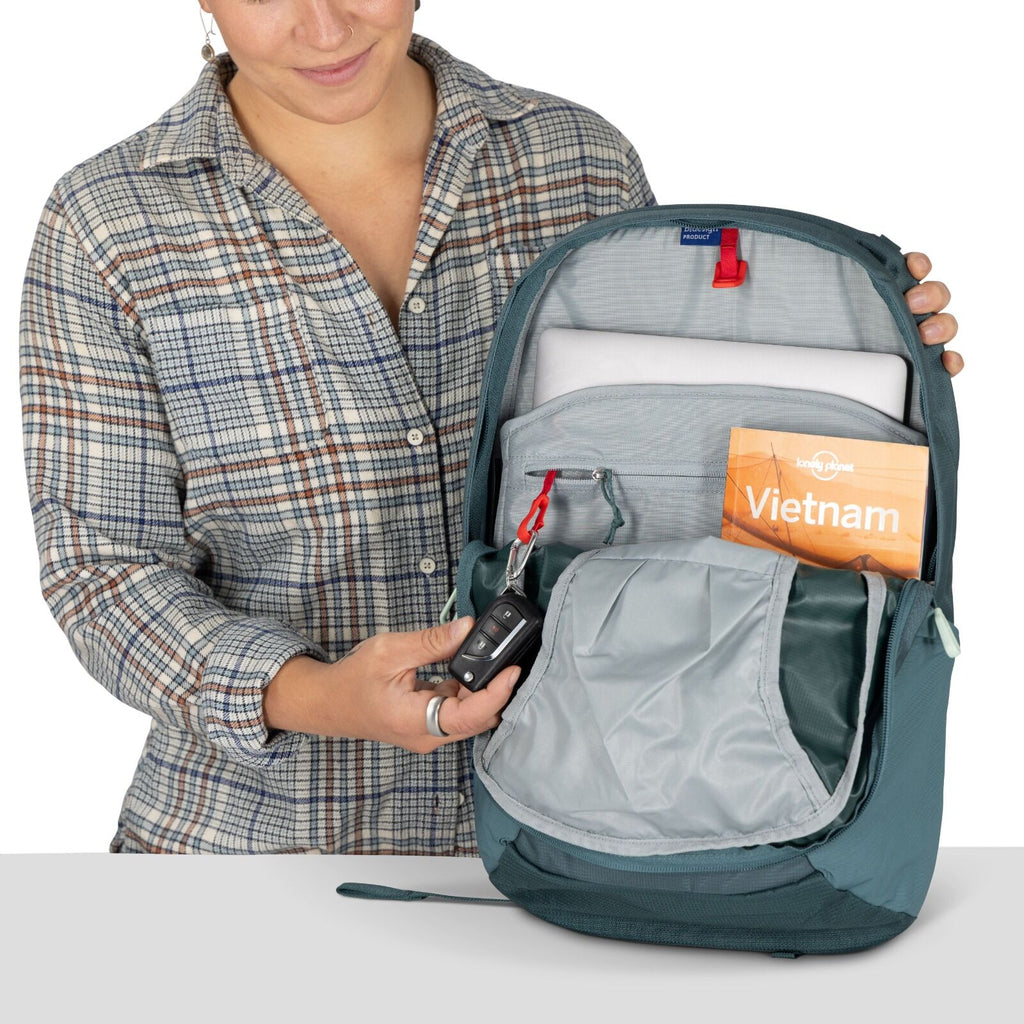 A person in a plaid shirt holds an open teal backpack. Inside, a guidebook titled "Vietnam," a laptop, and car keys can be seen. The scene suggests travel preparation.