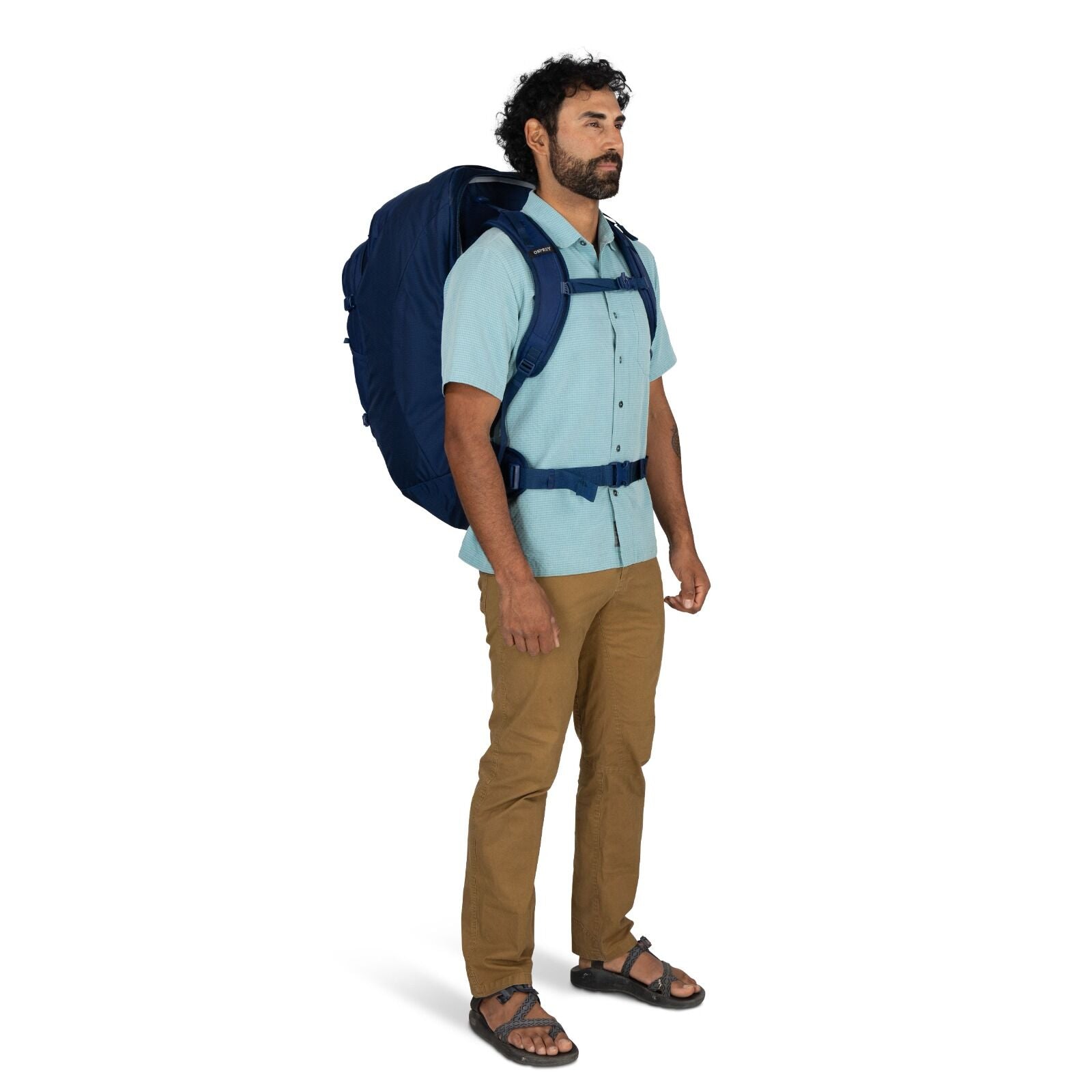 A man wearing a light blue short-sleeve shirt, khaki pants, and sandals stands with a large dark blue backpack on his back against a plain white background.