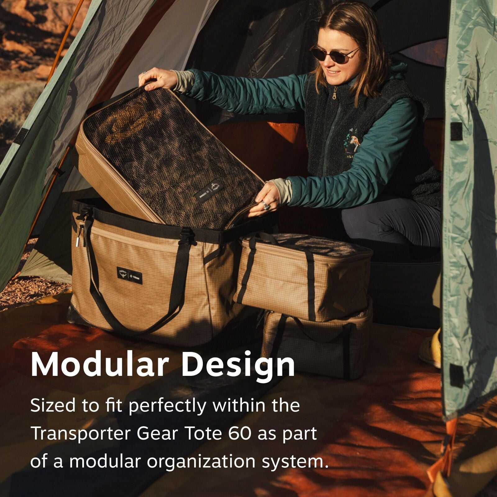 A person in a tent organizes camping gear using a modular storage system. They adjust a mesh-topped bag in a larger tote. Sunlight filters through trees.