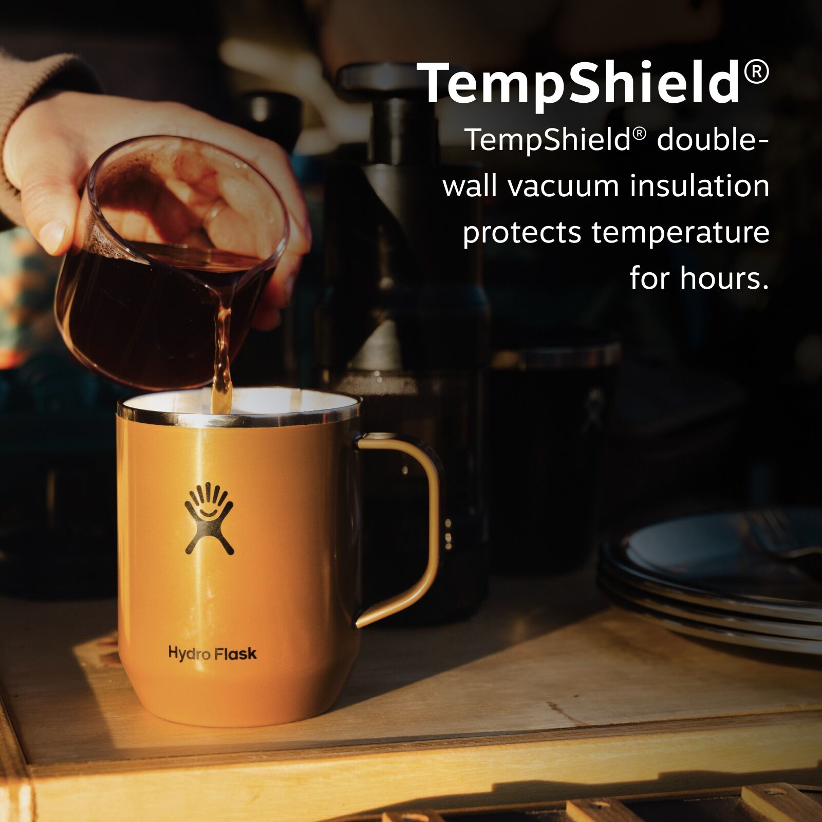 A hand pours coffee into a brown Hydro Flask mug on a table. Text reads, "TempShield® double-wall vacuum insulation protects temperature for hours." Cozy and inviting scene.