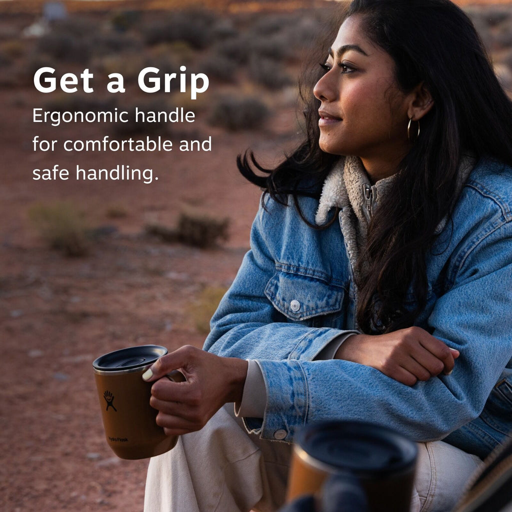 Woman in denim jacket sits outdoors, enjoying a drink from a brown mug with an ergonomic handle. Text reads "Get a Grip: Ergonomic handle for comfortable and safe handling." Background shows a desert landscape.
