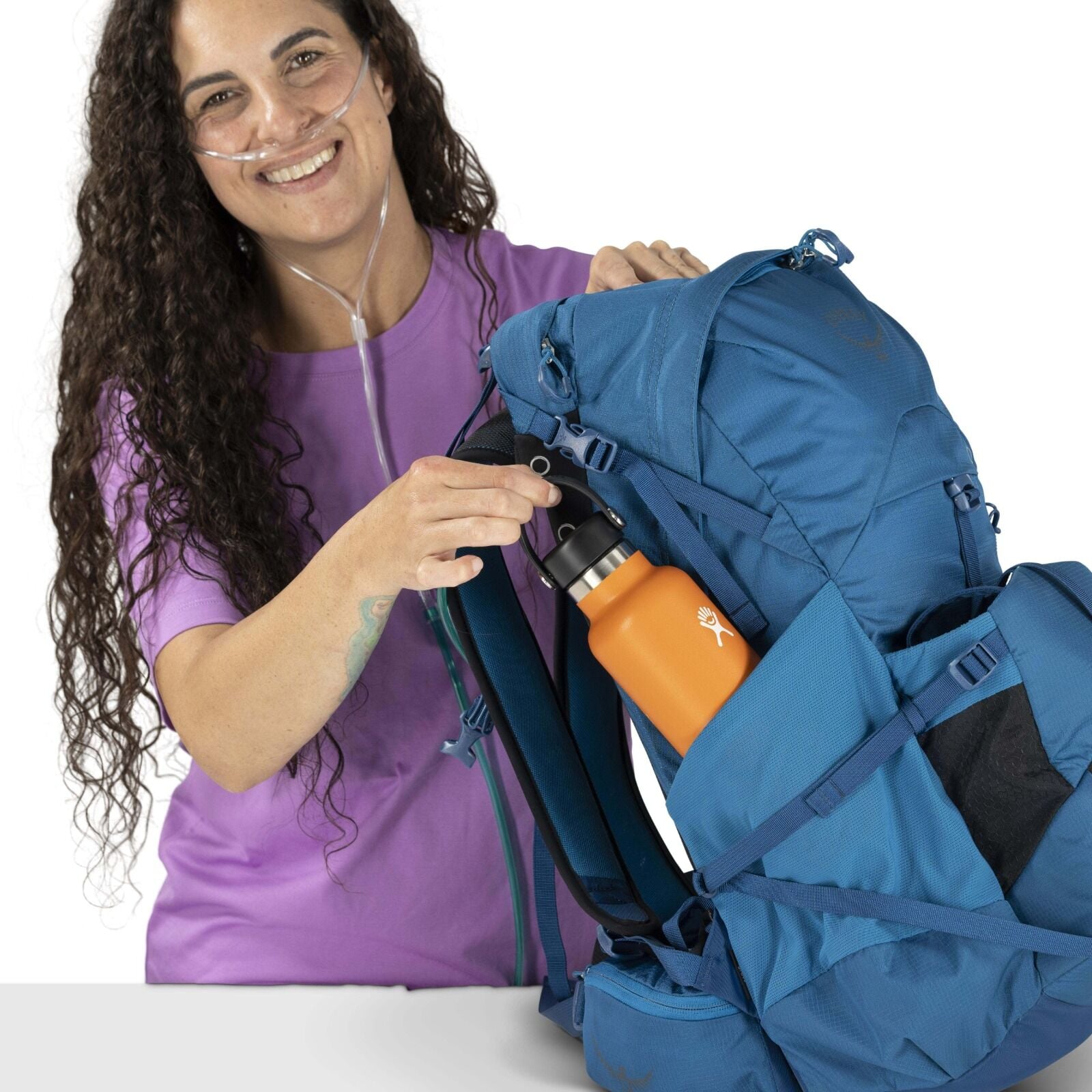 Woman in a purple shirt and oxygen tube, smiling and placing an orange water bottle into a large blue hiking backpack. Bright and cheerful tone.