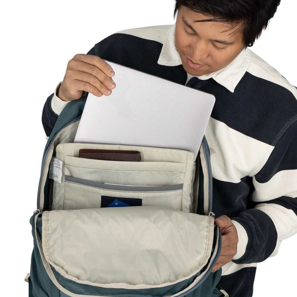 The image shows a person in a black and white striped long-sleeve shirt placing or removing a laptop from a backpack. The backpack is open, revealing an organized interior with various compartments, one of which holds the laptop. Additionally, there appea