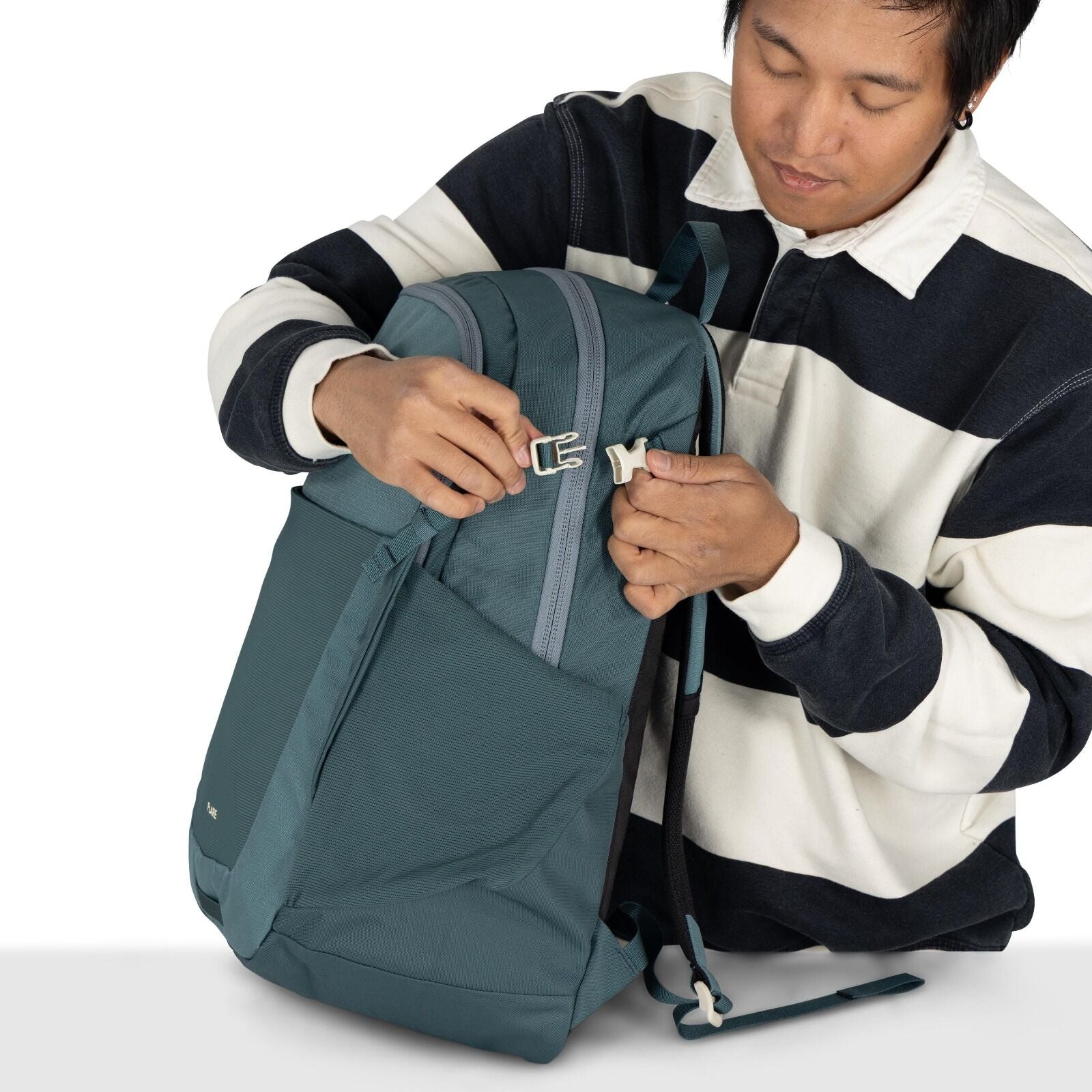 The image shows a person holding a teal-colored backpack, which they are in the process of securing by fastening a white buckle. The backpack has a sleek design with a large front pocket and a visible zipper on top. The person is wearing a black and white