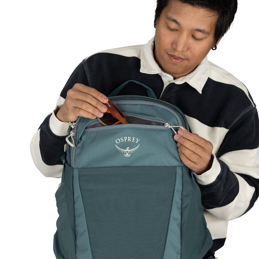 The image shows a person wearing a long-sleeve shirt with a thick horizontal striped pattern in black and white. They are holding a teal-colored backpack branded with the logo "Osprey" on the front. The backpack has multiple compartments, and the person i