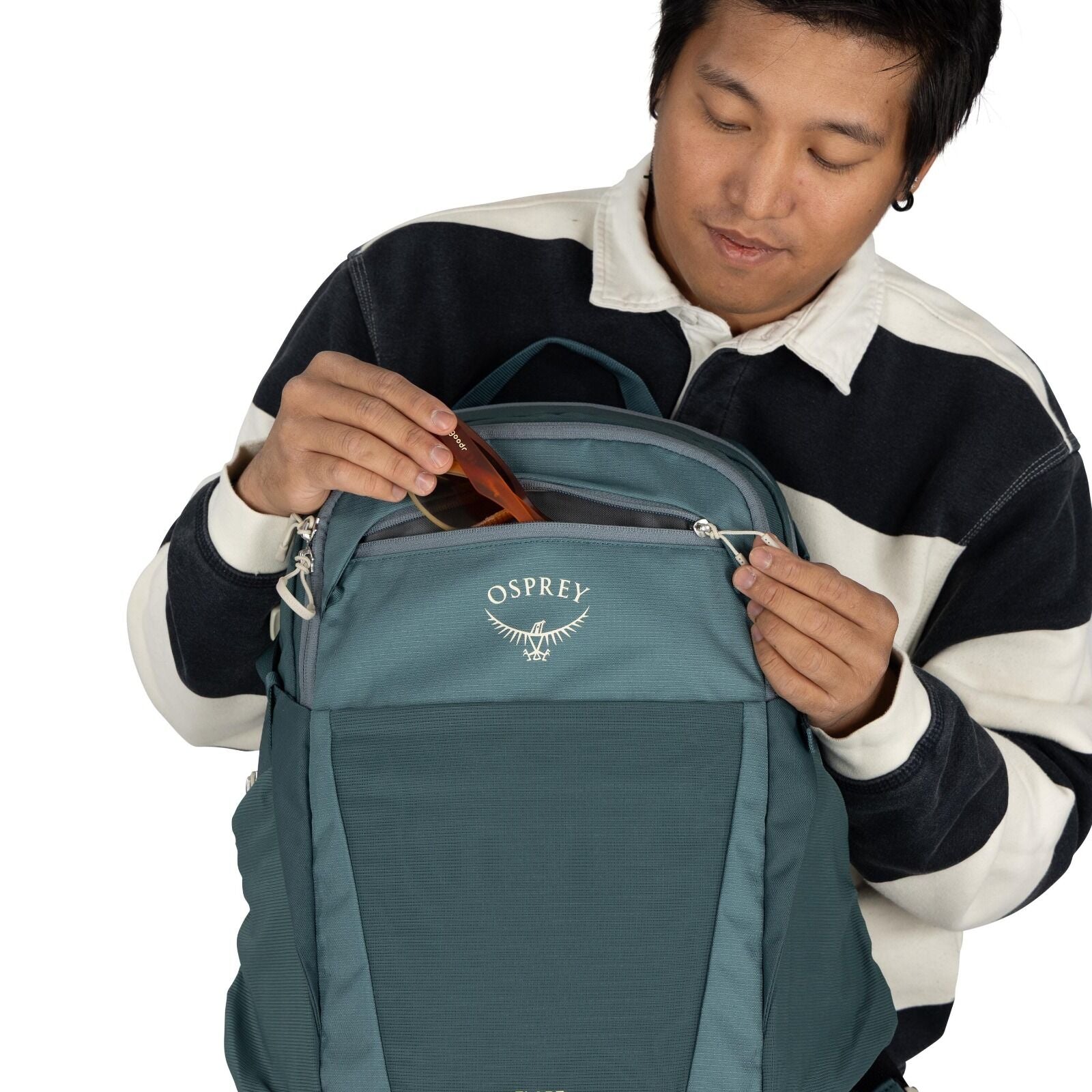 The image shows a person wearing a long-sleeve shirt with a thick horizontal striped pattern in black and white. They are holding a teal-colored backpack branded with the logo "Osprey" on the front. The backpack has multiple compartments, and the person i