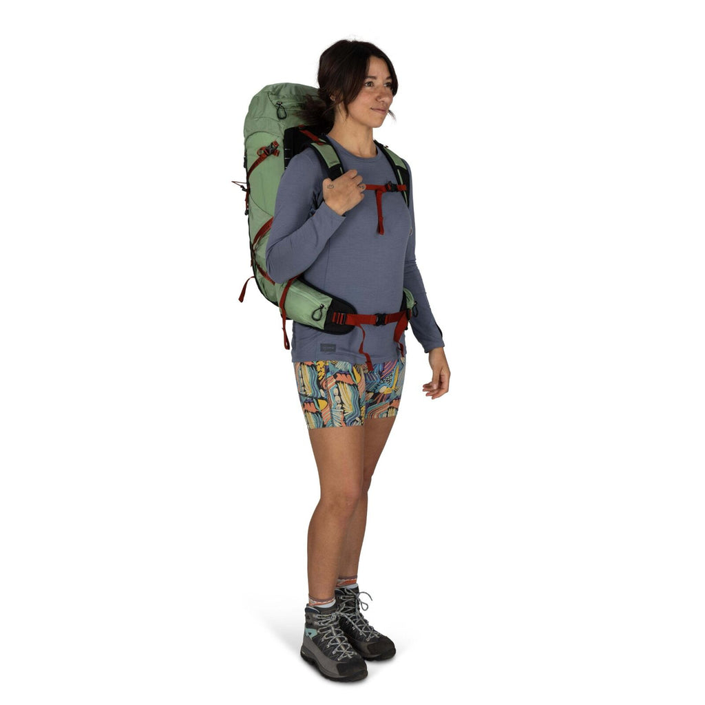A person stands with a large green backpack, wearing a long-sleeve shirt and colorful patterned shorts, ready for an outdoor adventure.