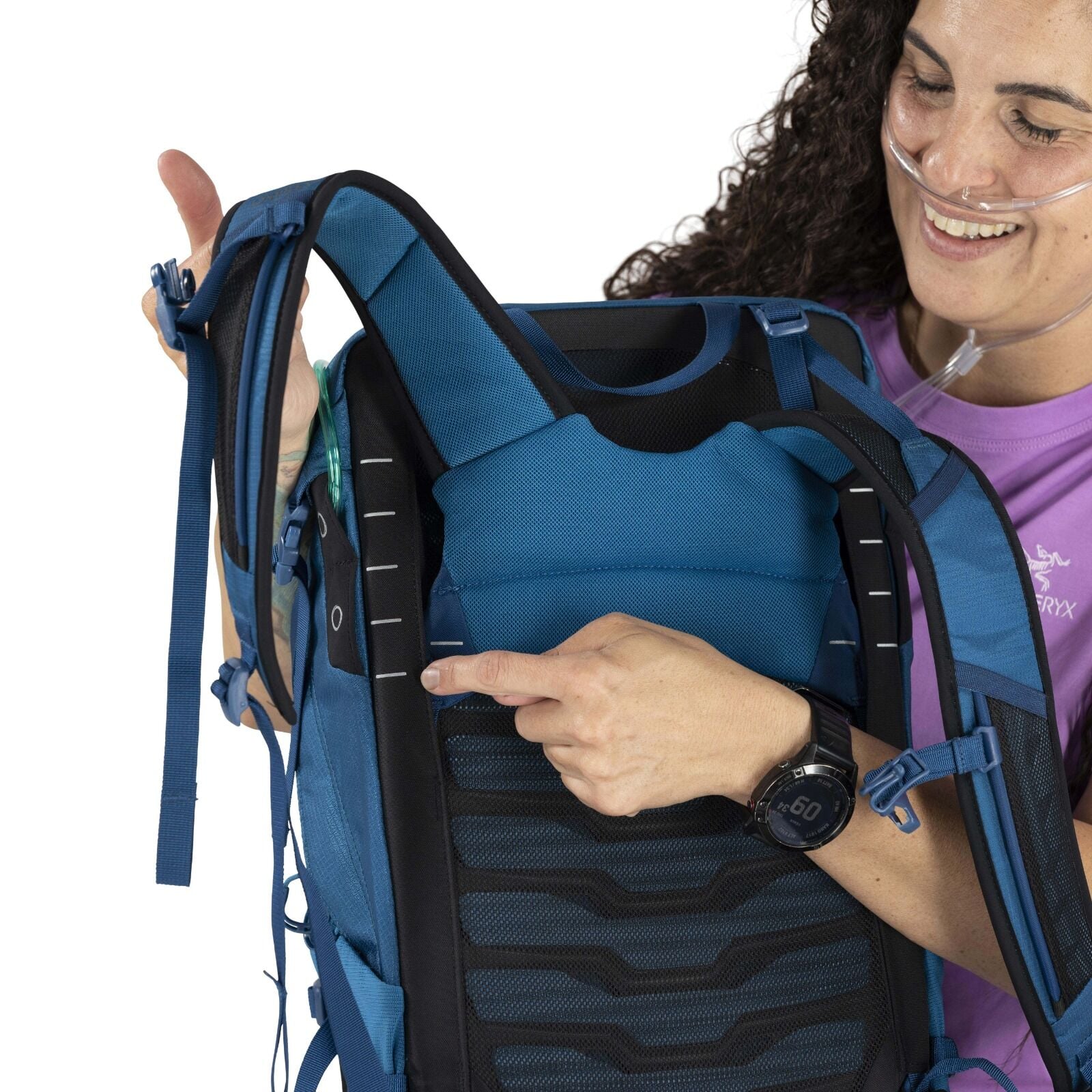 A woman with curly hair smiles while adjusting a blue backpack, pointing to its straps. She is wearing a purple shirt and an oxygen tube.