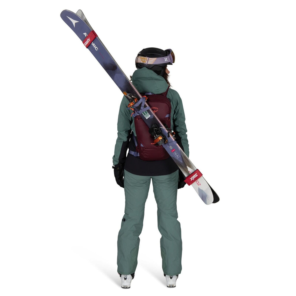 The image shows a person dressed in ski gear, viewed from the back. They are wearing a teal-colored ski jacket and pants, with a maroon backpack that securely holds a pair of skis diagonally across their back. The individual is also wearing black gloves a