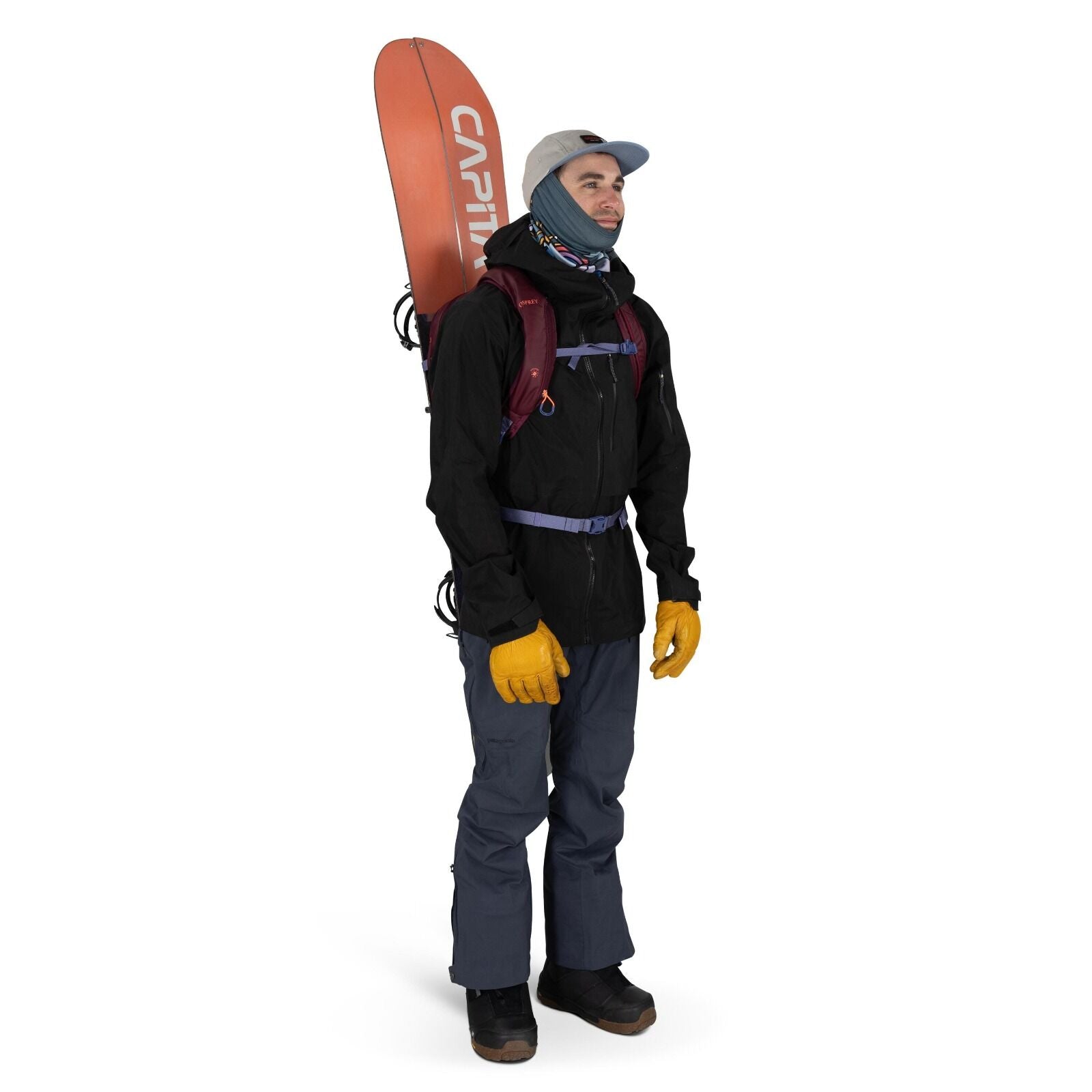 The image shows a person standing and dressed for snowboarding or outdoor winter sports. They are wearing a black jacket, blue-gray snow pants, and bright yellow gloves. A snowboard with the brand name "CAPiTA" written on it is secured on their back with