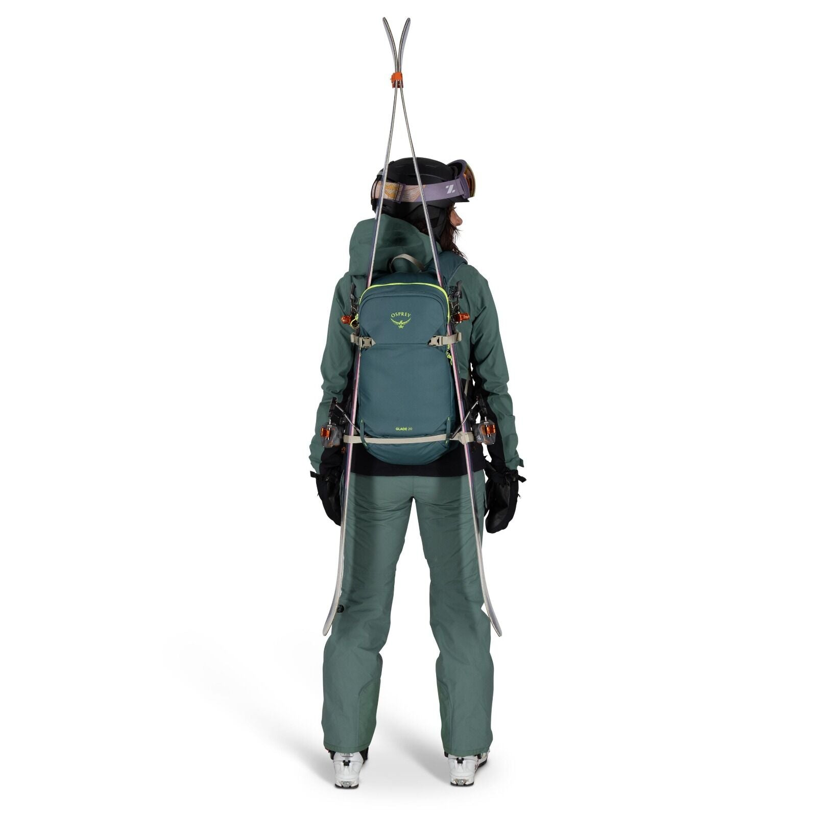 The image shows a person dressed in green winter attire standing with their back to the viewer. They are wearing a matching jacket and pants, along with gloves and ski boots, indicating they are prepared for a snowy outdoor adventure. The individual is ca