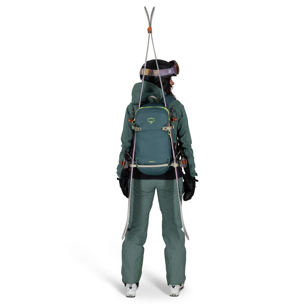 The image shows a person dressed in green winter attire standing with their back to the viewer. They are wearing a matching jacket and pants, along with gloves and ski boots, indicating they are prepared for a snowy outdoor adventure. The individual is ca
