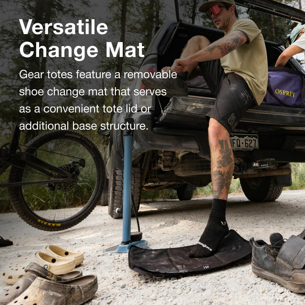 A person is using a removable shoe change mat next to an open truck, with a bicycle and casual footwear nearby in a forested setting.