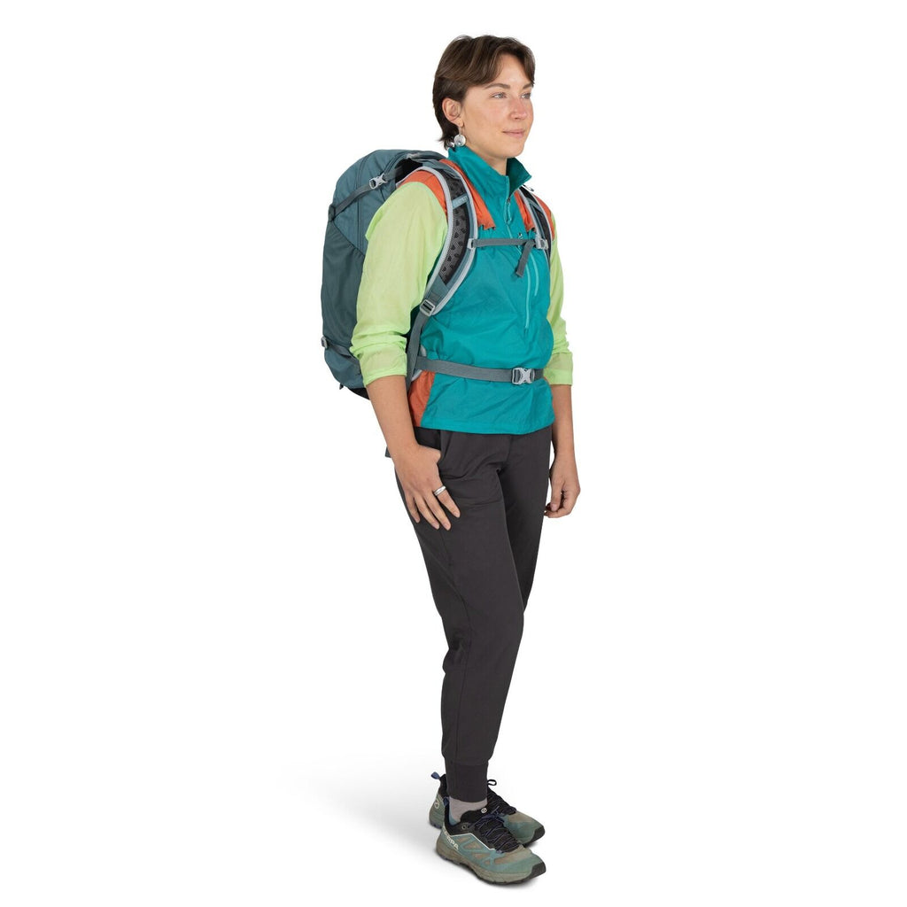 The image shows an individual wearing outdoor hiking attire, including a turquoise and orange jacket layered over a light green shirt. The person is also wearing dark gray pants and gray and teal hiking shoes. A large, dark teal hiking backpack is strappe
