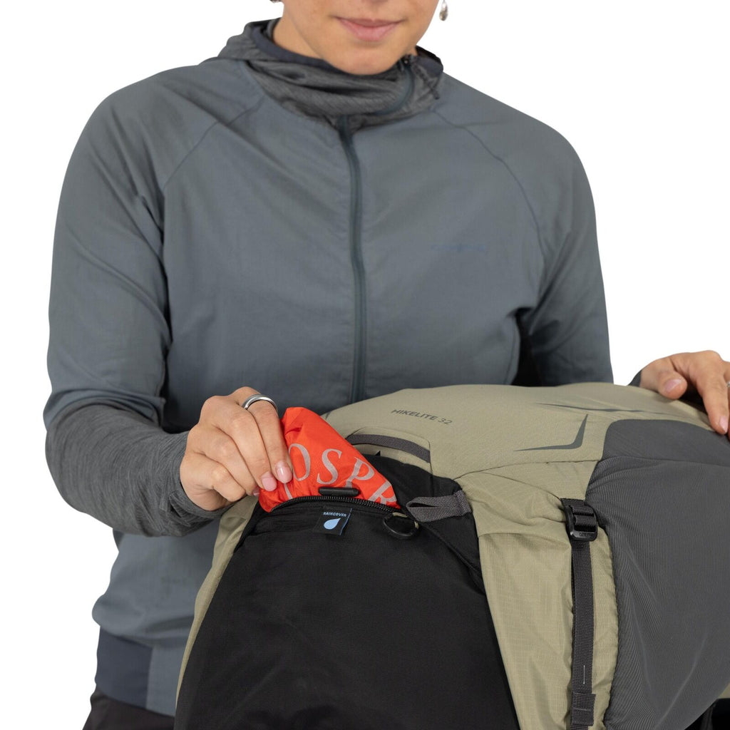 The image depicts a person wearing a gray jacket, reaching into a pocket of a green and black backpack. The backpack appears to be of the brand "Osprey," with the logo visible on the red fabric being pulled out of the pocket. The backpack has a prominent