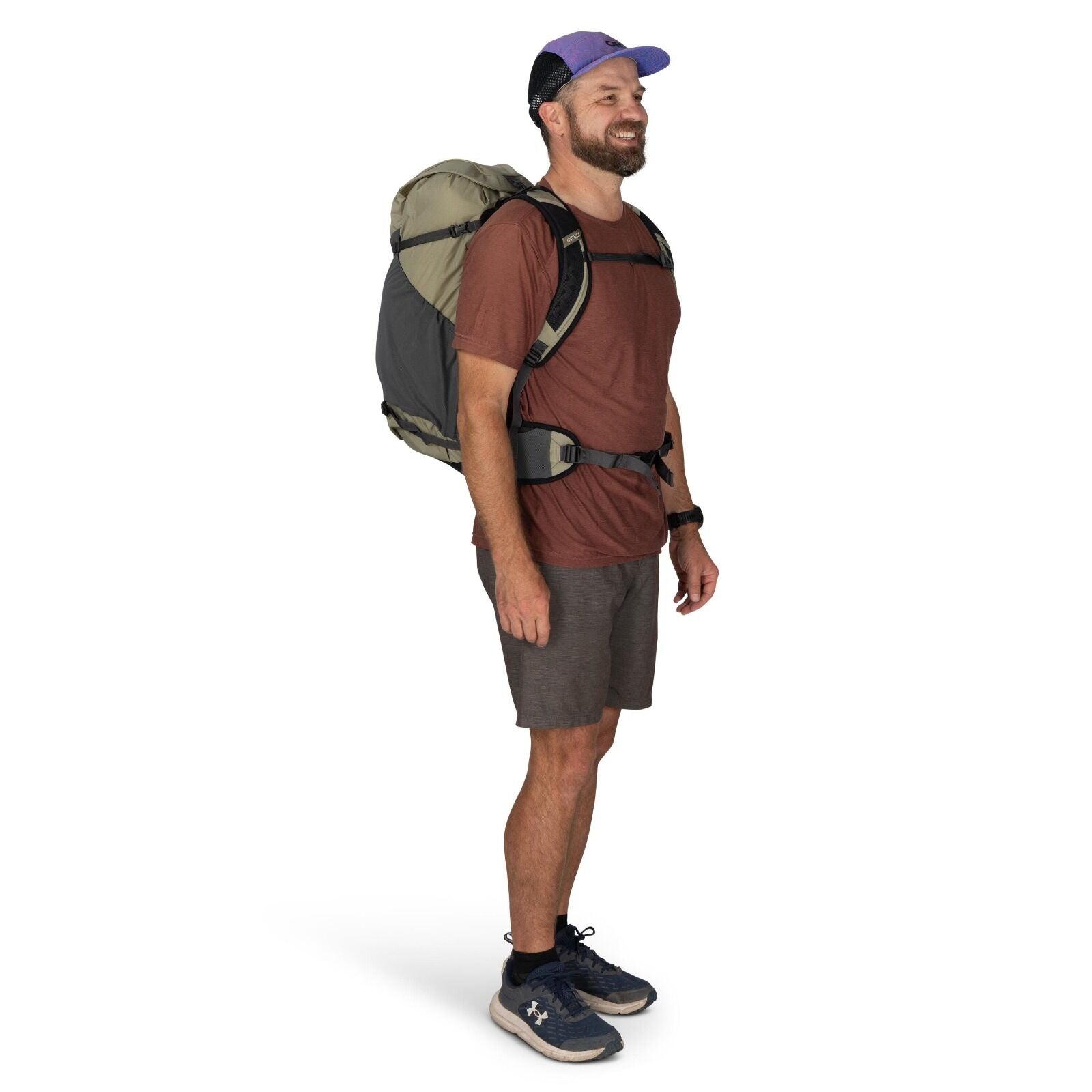 The image shows a person standing in a studio setting wearing casual outdoor attire. They are dressed in a reddish-brown t-shirt paired with dark gray shorts and navy blue athletic shoes. The person is carrying a large backpack, which is primarily green a