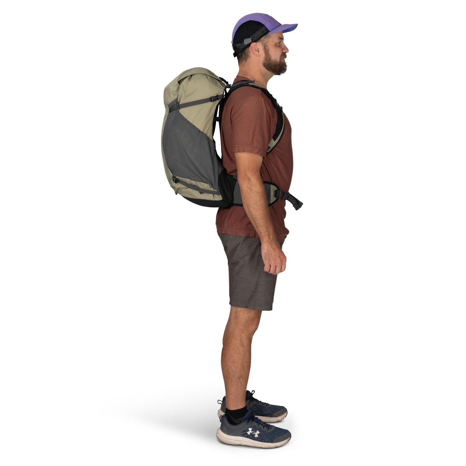 The image shows a man standing in profile against a plain white background. He is wearing a light brown t-shirt and dark brown shorts, along with blue athletic shoes with white soles that feature the Under Armour logo. On his back, he carries a large, gra