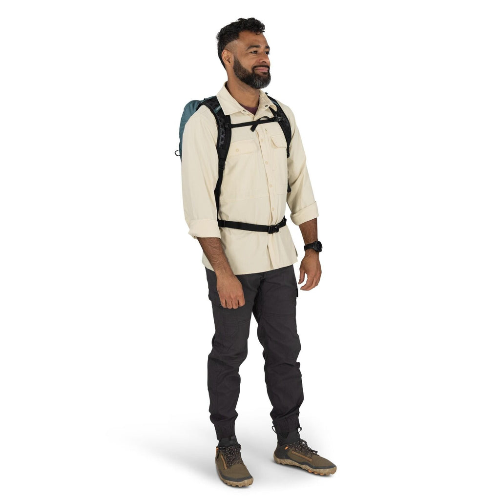 The image depicts a man standing in a studio setting, dressed in outdoor apparel suitable for hiking or trekking. He is wearing a light beige button-up long-sleeve shirt, which has been rolled up to the elbows, paired with dark grey cargo pants. On his fe