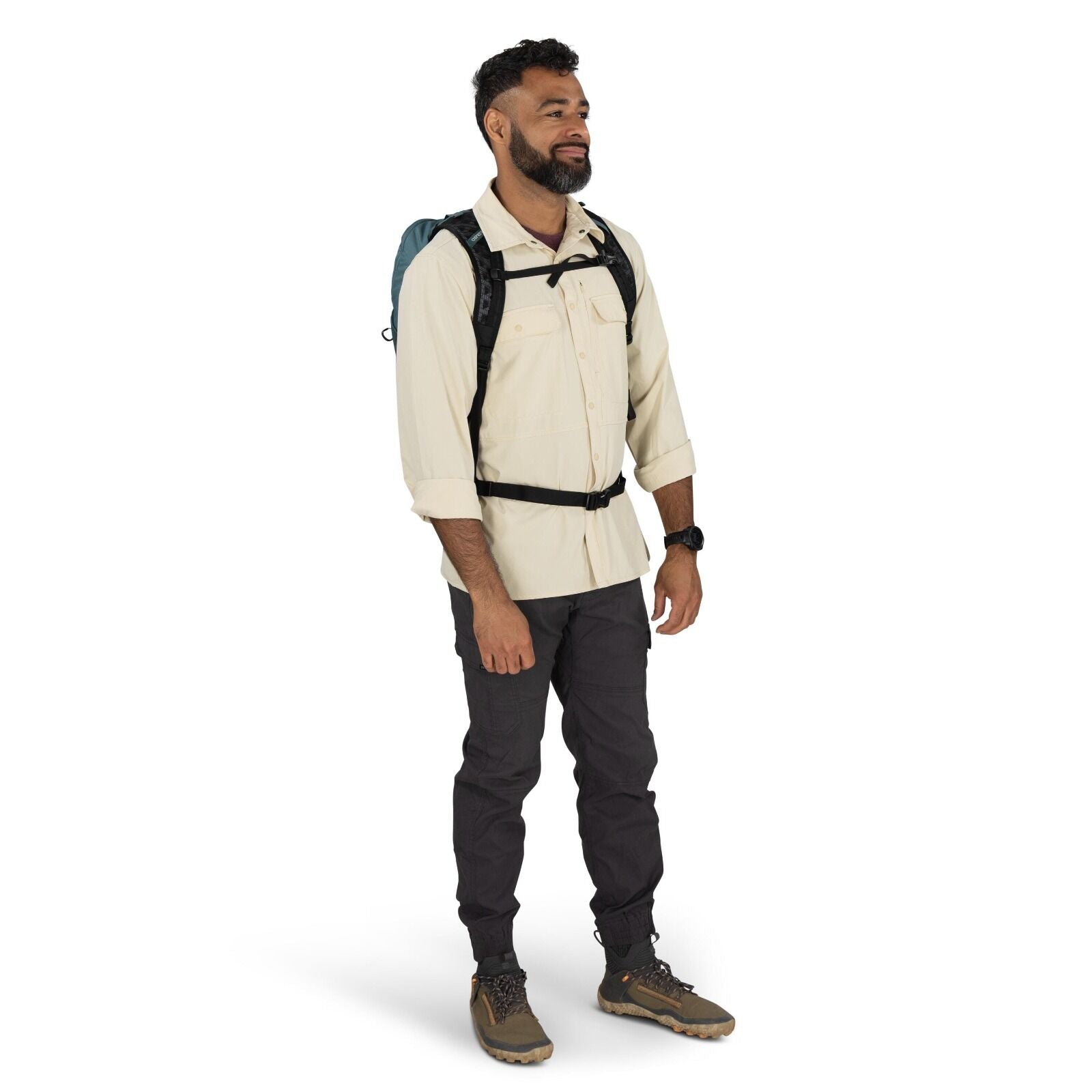 The image depicts a man standing in a studio setting, dressed in outdoor apparel suitable for hiking or trekking. He is wearing a light beige button-up long-sleeve shirt, which has been rolled up to the elbows, paired with dark grey cargo pants. On his fe