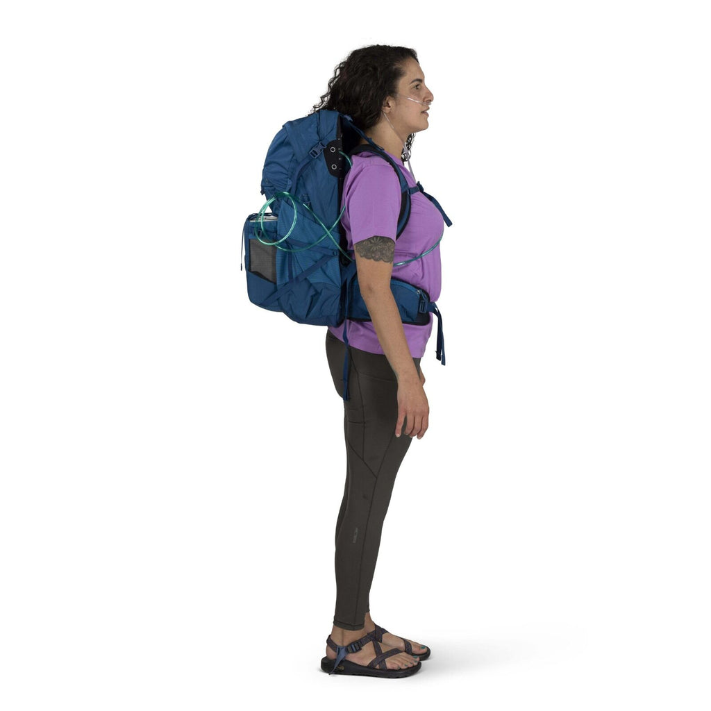 Side profile of a person wearing a blue hiking backpack, purple shirt, and dark leggings, smiling. They have shoulder-length curly hair and wear sandals.