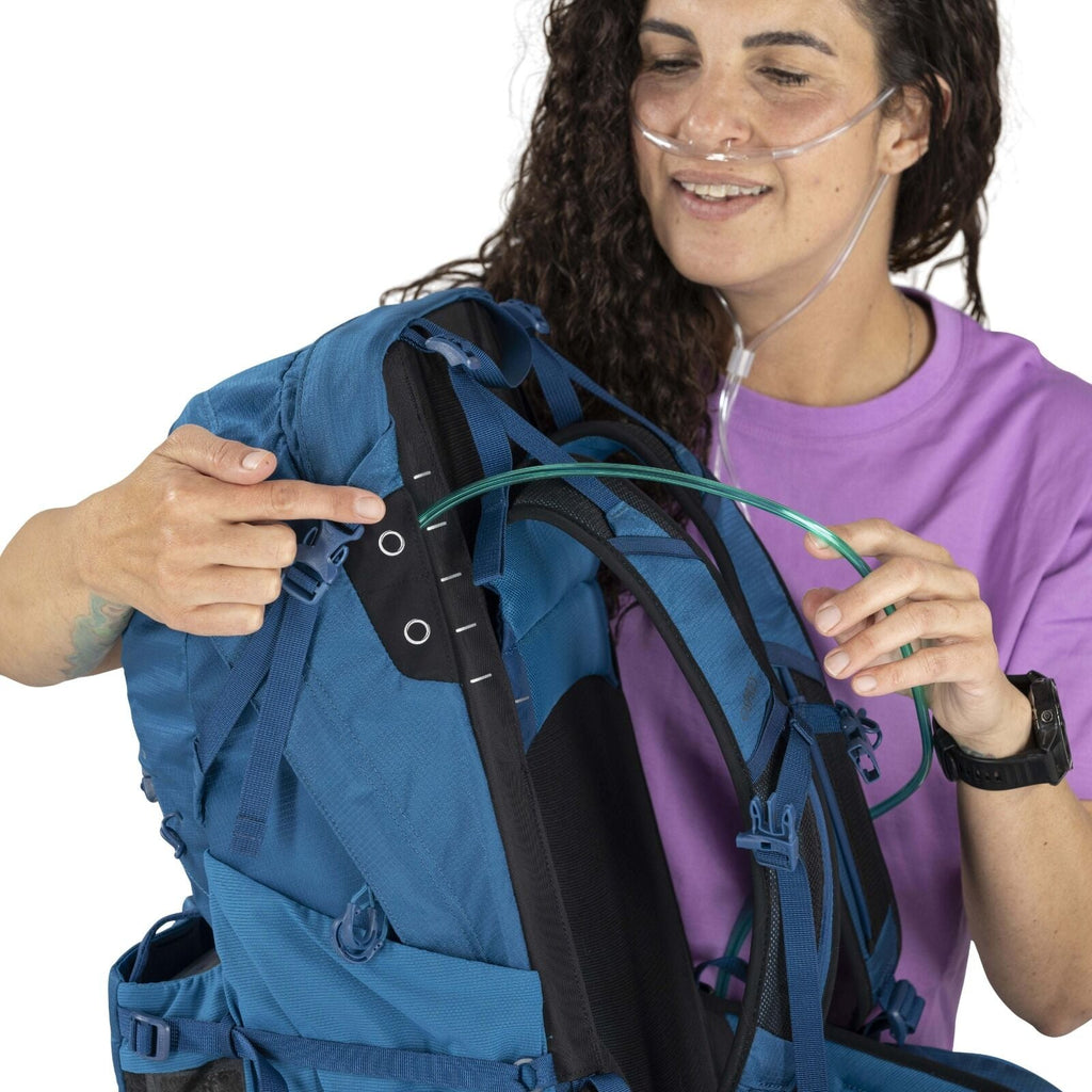 A person with curly hair and wearing an oxygen nasal cannula is holding and pointing to a blue backpack, highlighting its features with a smile.