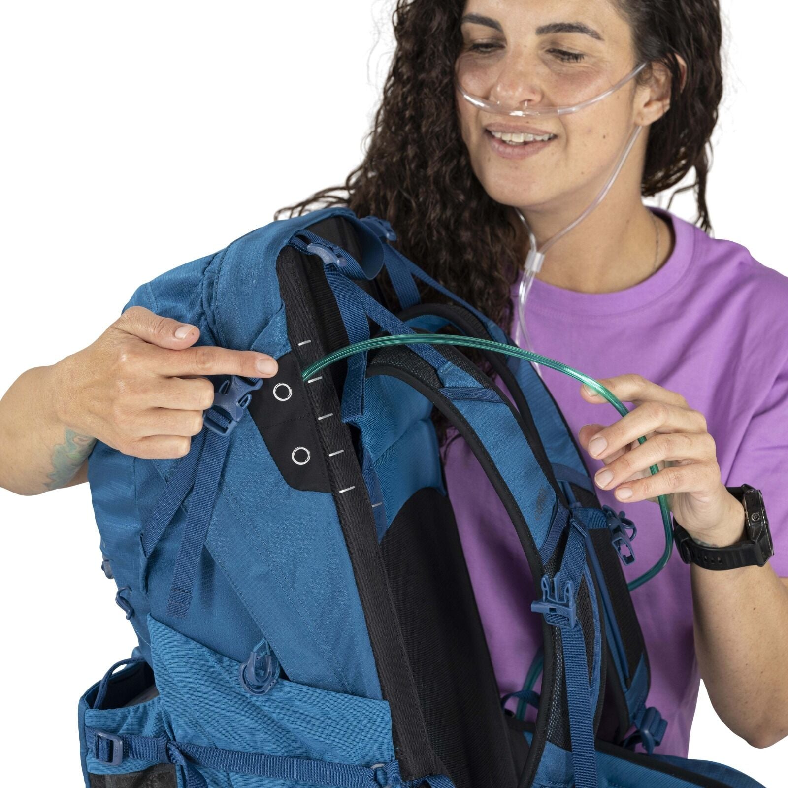 A person with curly hair and wearing an oxygen nasal cannula is holding and pointing to a blue backpack, highlighting its features with a smile.