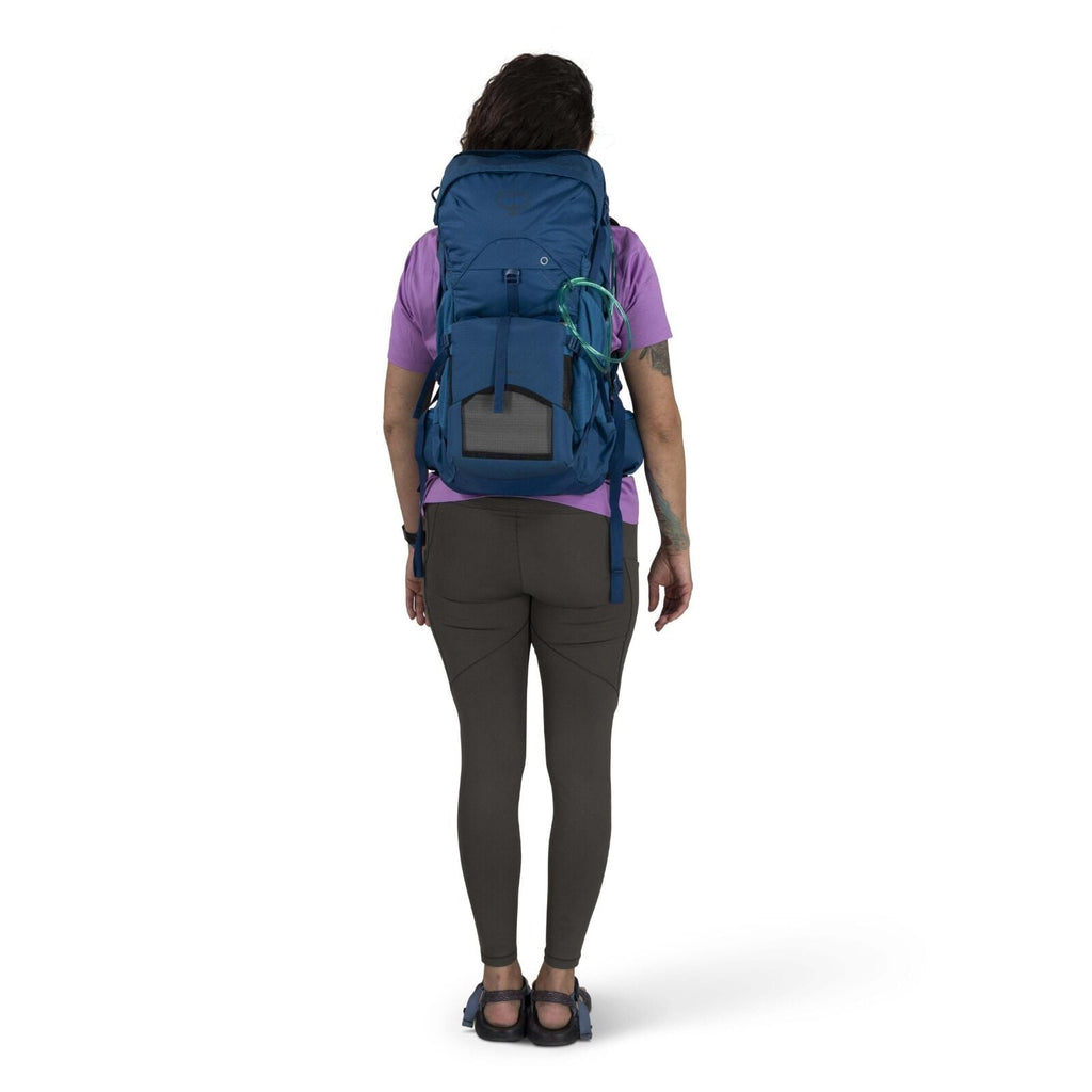 A person facing away, wearing a blue hiking backpack, purple shirt, and brown leggings. The background is plain white, emphasizing outdoor readiness.
