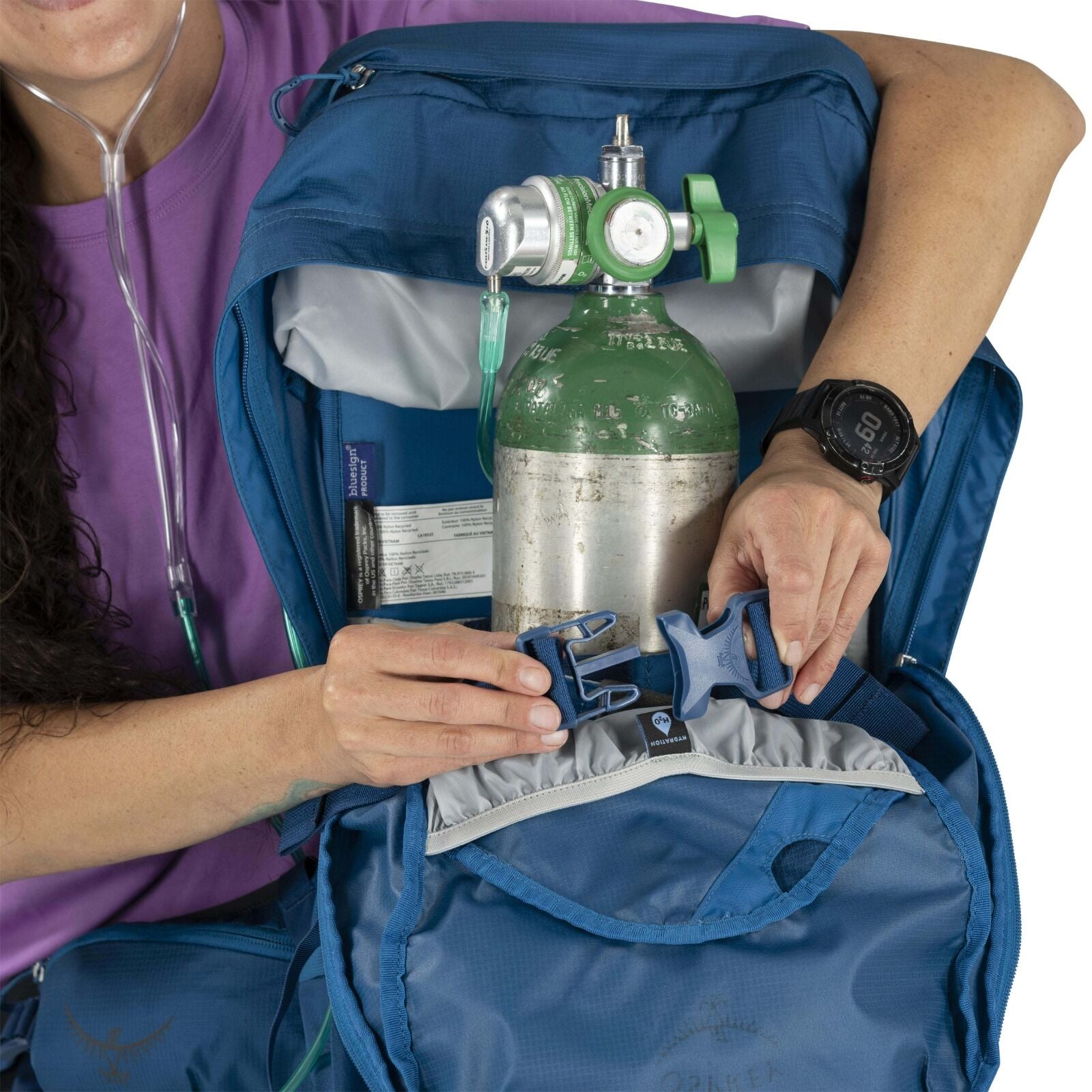 A person in a purple shirt is opening a blue backpack containing an oxygen tank. The setting suggests preparedness and medical care.