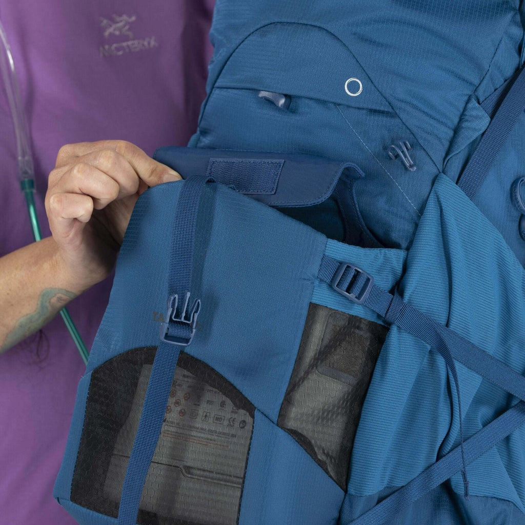 A person in a purple shirt adjusts the strap of a blue backpack with multiple compartments and mesh pockets, emphasizing functionality and detail.