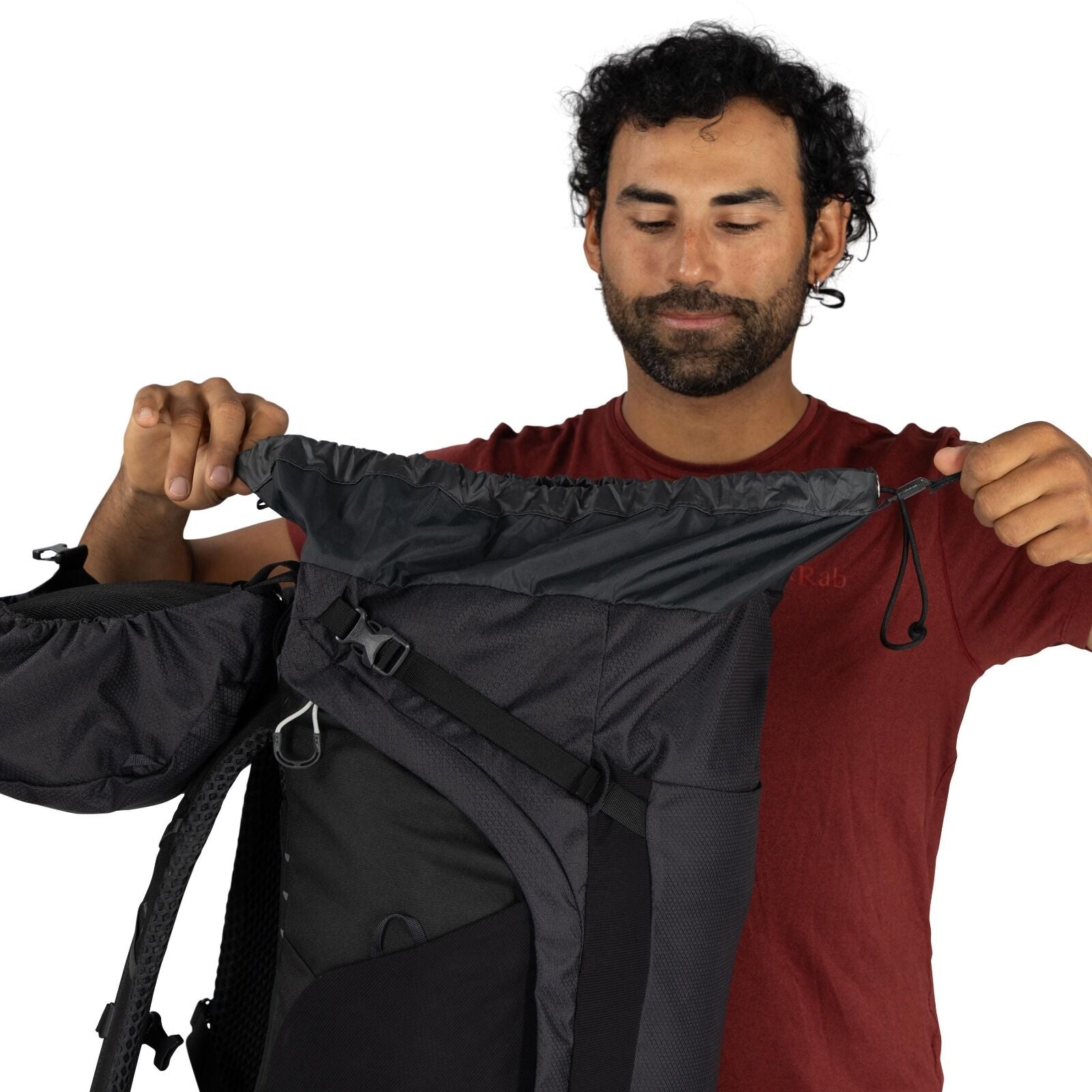 The image shows a person wearing a red Rab t-shirt, demonstrating the features of a black and gray backpack. They are holding the backpack open, highlighting the expandable drawstring closure at the top. The backpack appears to have multiple compartments
