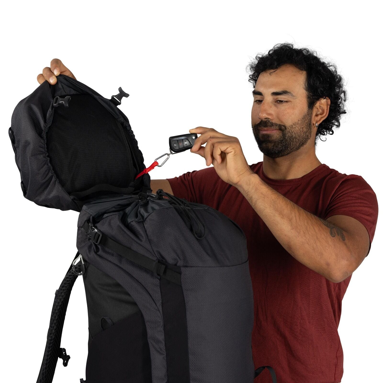 The image features a person wearing a short-sleeved maroon shirt, holding a large, black backpack. The individual is adjusting or examining something inside the open top compartment of the backpack. Attached to the zipper of the backpack is a red strap co
