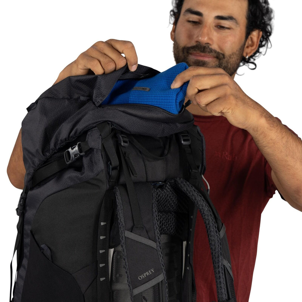 The image features a person wearing a red Rab t-shirt, who is adjusting or packing a black Osprey backpack. The individual is holding open the top of the backpack, revealing a blue internal compartment. The backpack is equipped with numerous straps and bu