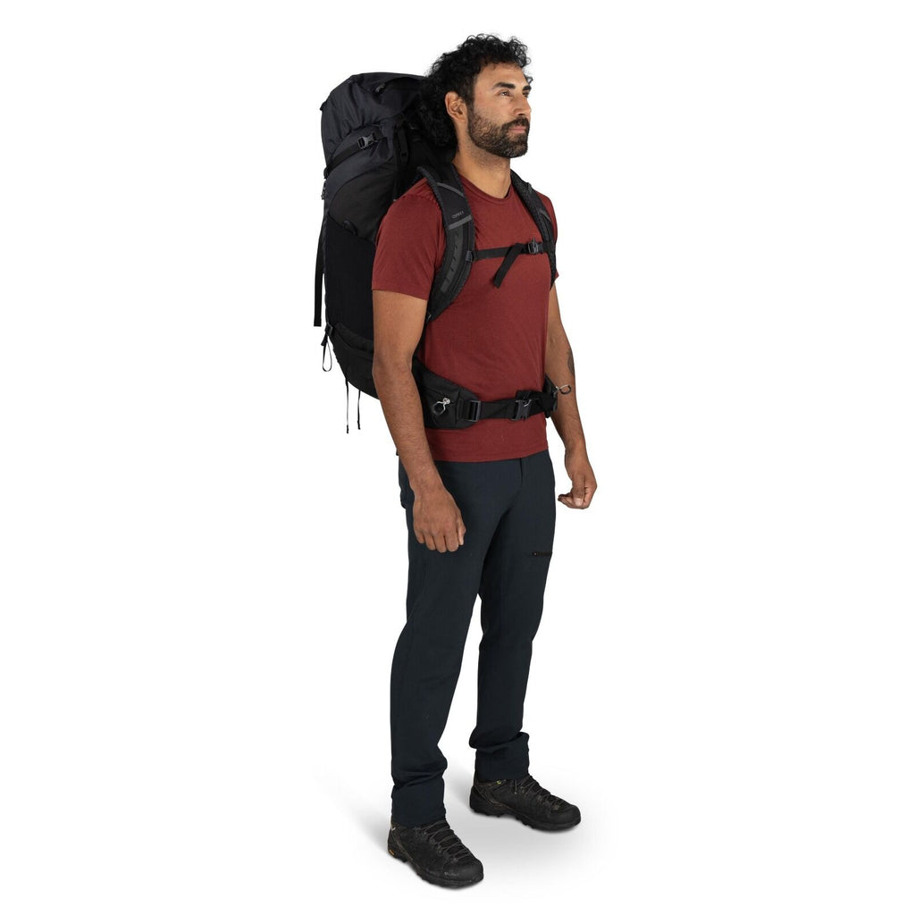 The image shows a person standing in profile view, dressed in hiking gear. They are wearing a red short-sleeved t-shirt and dark-colored hiking pants. The individual is carrying a large black backpack with various straps and attachments, suggesting it is