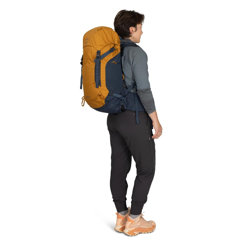 The image shows a person standing sideways, wearing outdoor clothing suitable for hiking or trekking. They are equipped with a large yellow and dark blue backpack labeled "ATLAS," which features multiple straps and adjustments for secure fitting. The pers